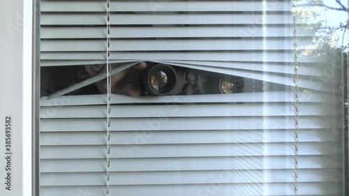 A man is observing with binoculars through the blinds in the window.