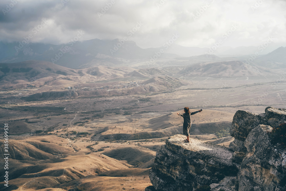 Woman standing on rock high in mountains and watches landscape of hills.