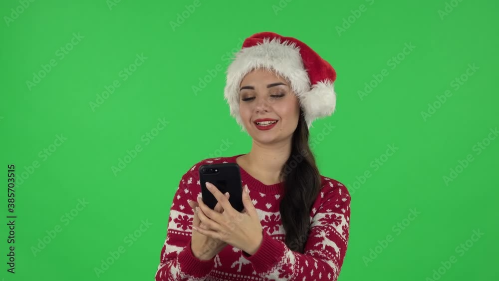 Portrait of sweety girl in Santa Claus hat is making selfie on mobile phone then looking photos. Green screen Portrait of sweety girl in Santa Claus hat is making selfie on mobile phone then looking photos. Green screen