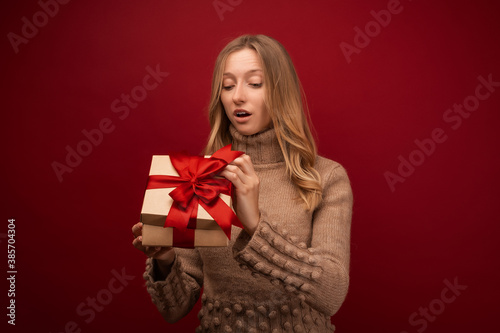 Charming blonde woman open a gift with red ribbon. Studio shot red background. New Year Birthday Holiday concept