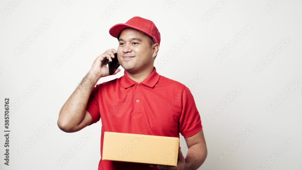 Smiling young asian delivery man in red uniform holding parcel cardboard box using smartphone talking with customer on isolated white background. 4k Resolution. Smiling young asian delivery man in red uniform holding parcel cardboard box using smartphone talking with customer on isolated white background. 4k Resolution.