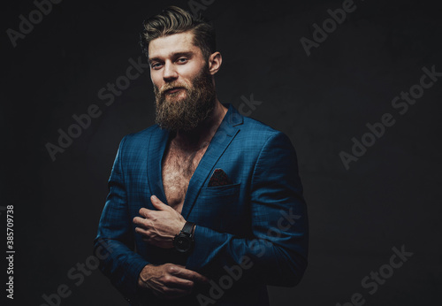 Styled and handsome businessman in custom blue suite with beard and modern haircut posing holding his jacket sleeve in dark background.