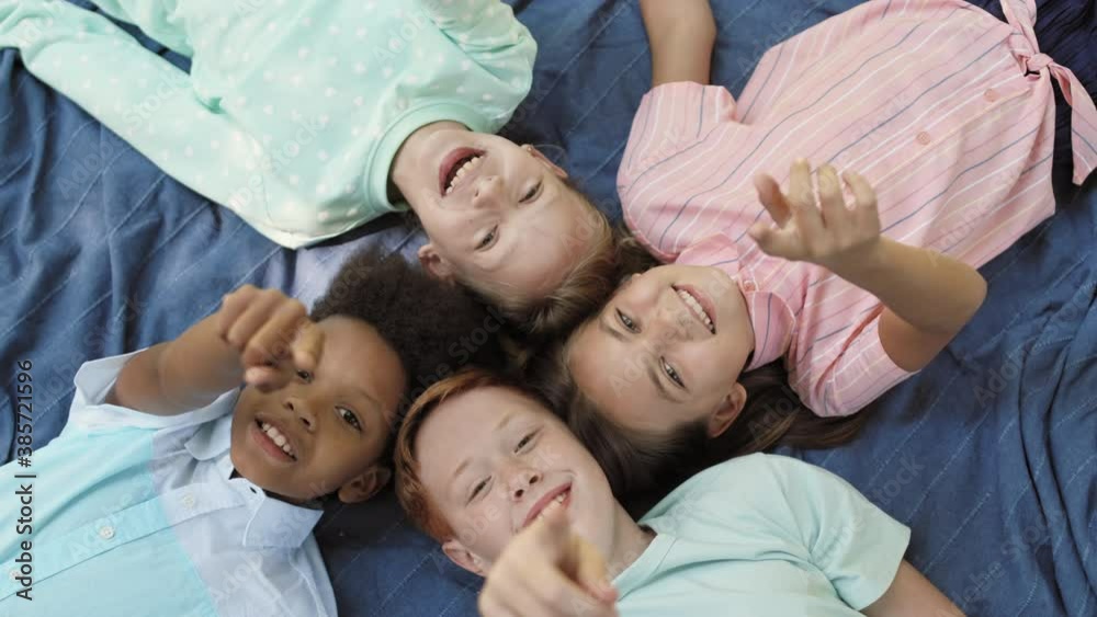 Top close-up of four Caucasian and African school boys and girls laying down on blue blanket head to head, laughing looking and pointing fingers on camera