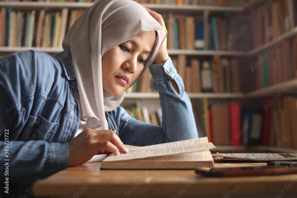 Asian muslim female student, studying hard in library, reading books, preparing for exam, tired exhausted expression