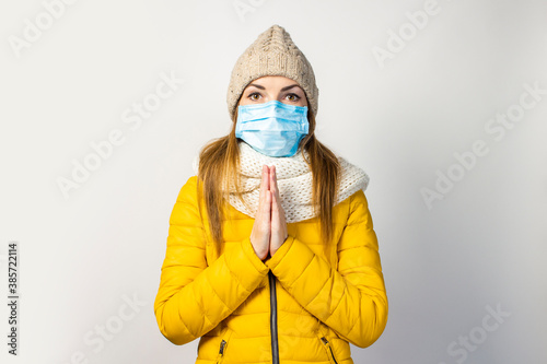 young girl in a yellow jacket and hat, a medical mask on her face, holds her hands with palms together, praying, on a light background. Concept of hope, virus, infectious disease