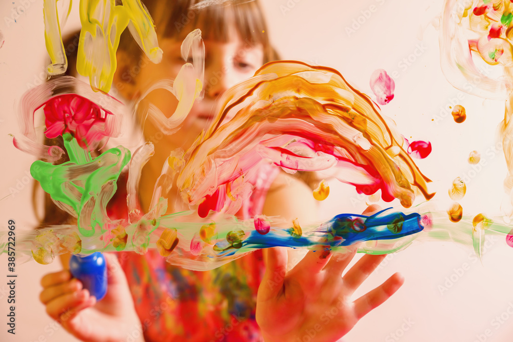Obraz premium Beautiful young girl painting artwork on glass with colorful fingers. Happy childhood, art, painting lessons concept. Selective focus on finger.