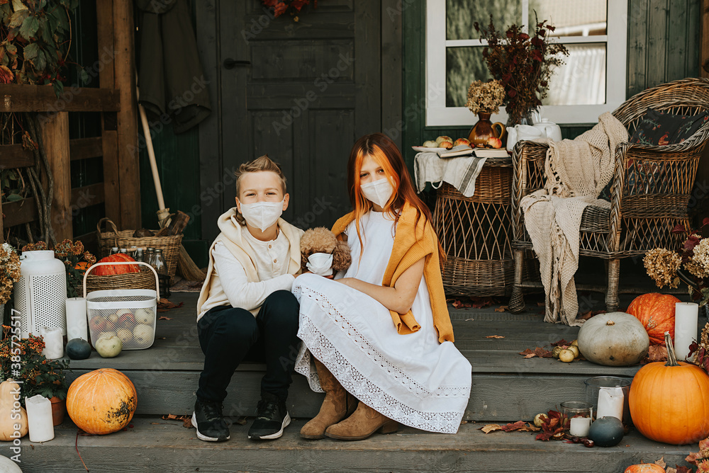 children a boy and a girl with their poodle dog in protective masks on the porch of the backyard decorated with pumpkins in autumn, the concept of the covid-19 pandemic quarantine