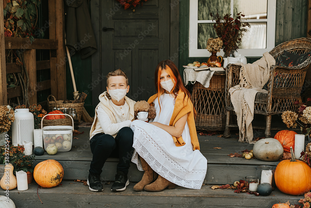 Fototapeta premium children a boy and a girl with their poodle dog in protective masks on the porch of the backyard decorated with pumpkins in autumn, the concept of the covid-19 pandemic quarantine