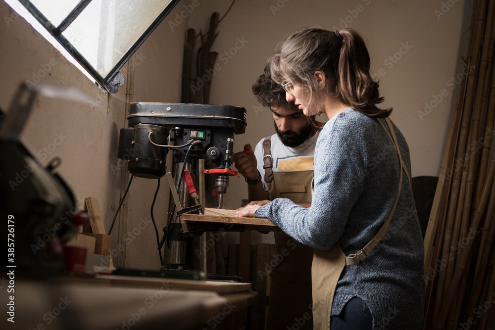 A young couple of carpenter working together in a small wood laboratory using a drill machine for timber. Couple crafting new home furniture in a carpentry workshop. young entrepreneurs concept