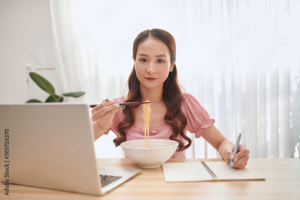 Young Asian woman eating noodles and working with laptop at home. Young Asian woman eating noodles and working with laptop at home.