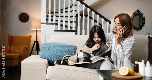 Beautiful Caucasian woman speaking on cellphone while sitting on couch in room with little sick daughter. Mother talking with doctor on smartphone. Unwell small girl coughing at home. Virus concept