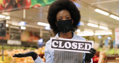 Close up portrait of African American beautiful female grocery store employee in mask and gloves standing in supermarket and holding sign Closed in quarantine. Woman seller indoors. Food store concept