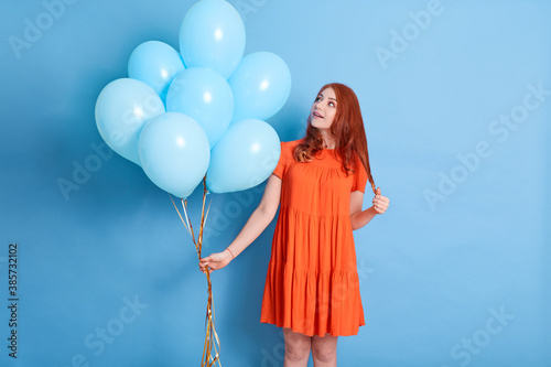 Woman holding bunch of helium balloons, looking at her present with surprised and excited facial expression, standing isolated over blue background. © sementsova321