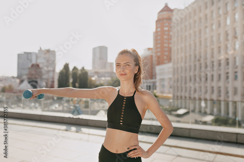 Happy exited woman in sportswear with hand dumbbells doing outside exercise in the city in sunny day