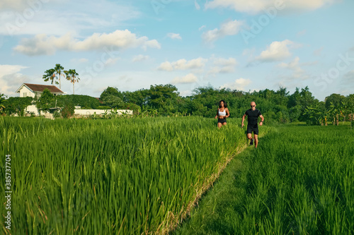 Mature Couple Running On Rice Field In Morning. Caucasian Man And Asian Woman On Jogging Workout Among Green Grass Against Tropical Landscape.
