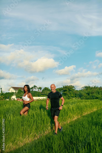 Mature Couple Running On Rice Field In Morning. Caucasian Man And Asian Woman On Jogging Workout Among Green Grass Against Tropical Landscape.