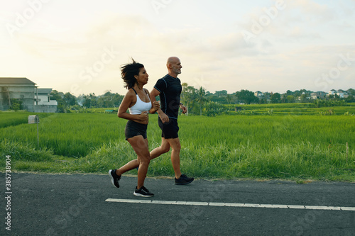 Mature Couple Running On Road At Tropical Landscape. Caucasian Man And Asian Woman On Jogging Workout In Morning Near Green Rice Fields.