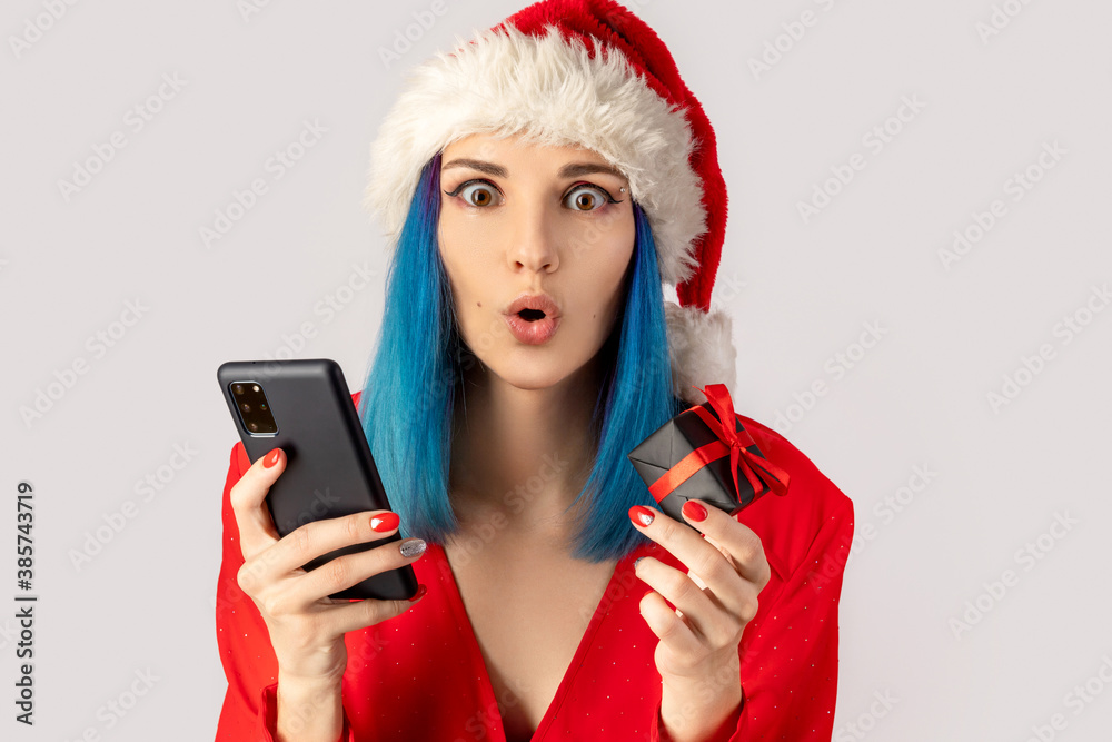 Naklejka premium Happy excited young woman in santa hat with gift box and smartphone