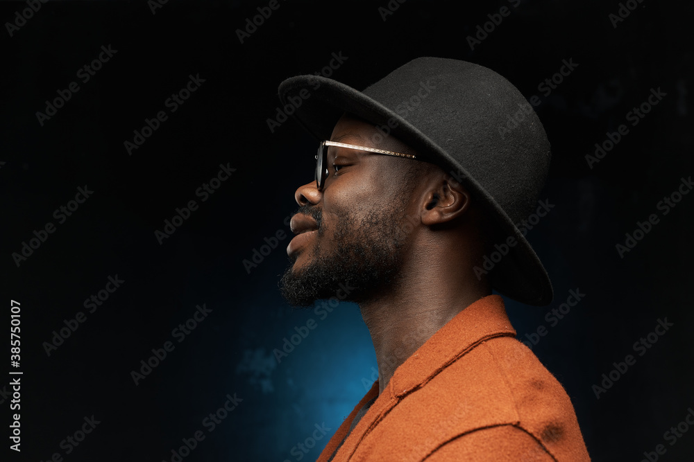profile photo of a black man in a black hat, brown jacket and jacket on a dark background in the studio, he is talking