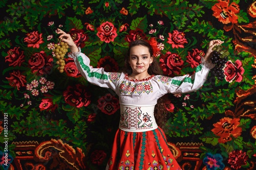 pretty moldavian girl in folk dress with grape racings in her hands. close up portrait. national traditional carpet on the background