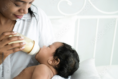 Happy African American baby infant drinking milk form bottle on mother arm. Breastfeeding. Portrait of a cute little baby sucking milk form bottle after wake up happily on mother's arms at home,