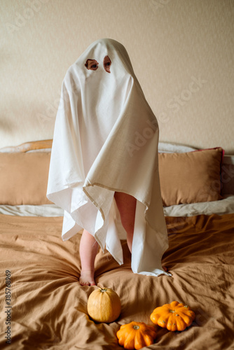 little child in a white sheet jumps on the bed at home, scares and plays ghost, halloween holiday