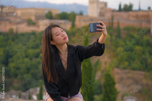 lifestyle portrait of young beautiful and happy Asian Chinese tourist woman having Summer holidays in Europe taking selfie with mobile phone at beautiful Alhambra of Granada in Spain