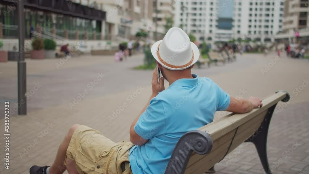 Male tourist in a white hat is resting while sitting on a bench, camera movement Male tourist in a white hat is resting while sitting on a bench, camera movement