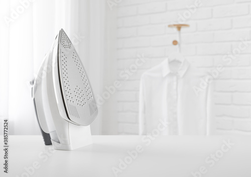 Photo Electric iron on table in blurred room with clothes rack