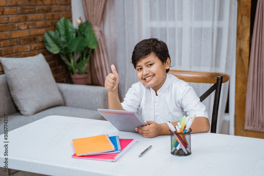 smiling asian boy holding tablet with thumbs up when sitting on a desk smiling asian boy holding tablet with thumbs up when sitting on a desk