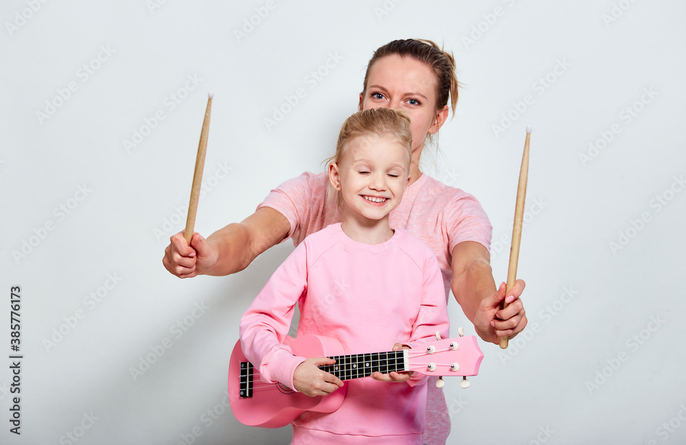 Yong mother and her pretty daughter playing on musical instruments, neutral gray background. Spending funny time together holding ukulele and wood drumsticks. Yong mother and her pretty daughter playing on musical instruments, neutral gray background. Spending funny time together holding ukulele and wood drumsticks.