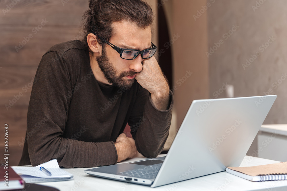 A serious, focused man is thinking while sitting at his desk at home and working on his laptop. A serious, focused man is thinking while sitting at his desk at home and working on his laptop.