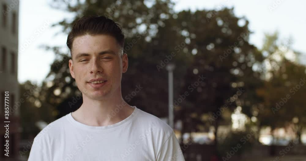 Portrait of young caucasian man brunette friendly looking to camera. Close up of millennial teen guy in white shirt clothes smiling while standing posing at street outdoors. Concept of people emotions