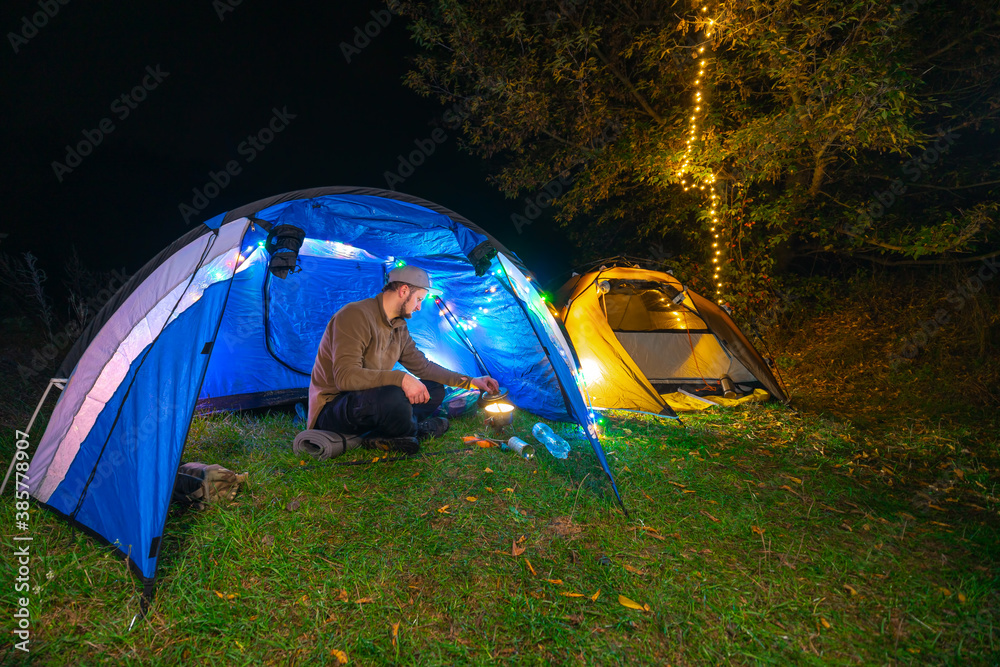 The guy prepares food, a mini gas burner. A pot and a bottle of water. Overnight in a tent. Camping, A man sits inside. The light of the gerlands. Night. Leisure and hobbies. Hiking. Copy space