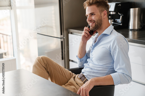Handsome joyful guy smiling and talking on mobile phone while sitting