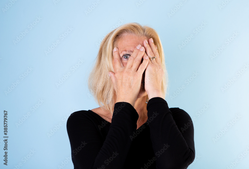 Studio portrait of 60+ woman with blonde page looking through her fingers on light blue background. Studio portrait of 60+ woman with blonde page looking through her fingers on light blue background.