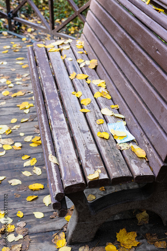 old wooden bench with dropped sanitary face mask covered by yellow fallen leaves in city park on autumn day