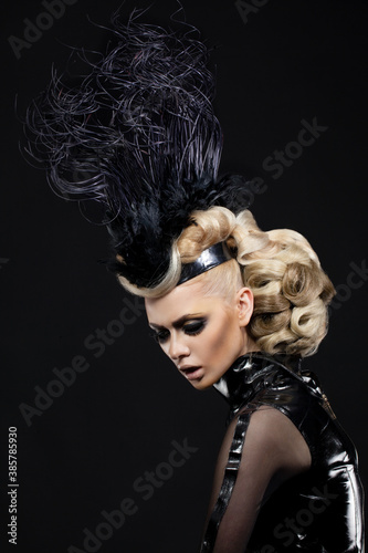 Blonde woman with artistic hairstyle and makeup, in sexy black clothes and hat with feather posing on black background.