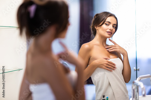 Smiling young woman in bathrobe and towel on head looking at mirror in bathroom