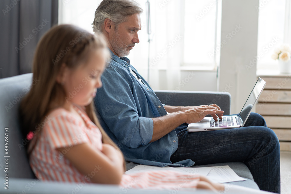No Time For Child. Grey-haired dad busy with laptop, working online at home, sad bored offended daughter sitting near by. No Time For Child. Grey-haired dad busy with laptop, working online at home, sad bored offended daughter sitting near by.