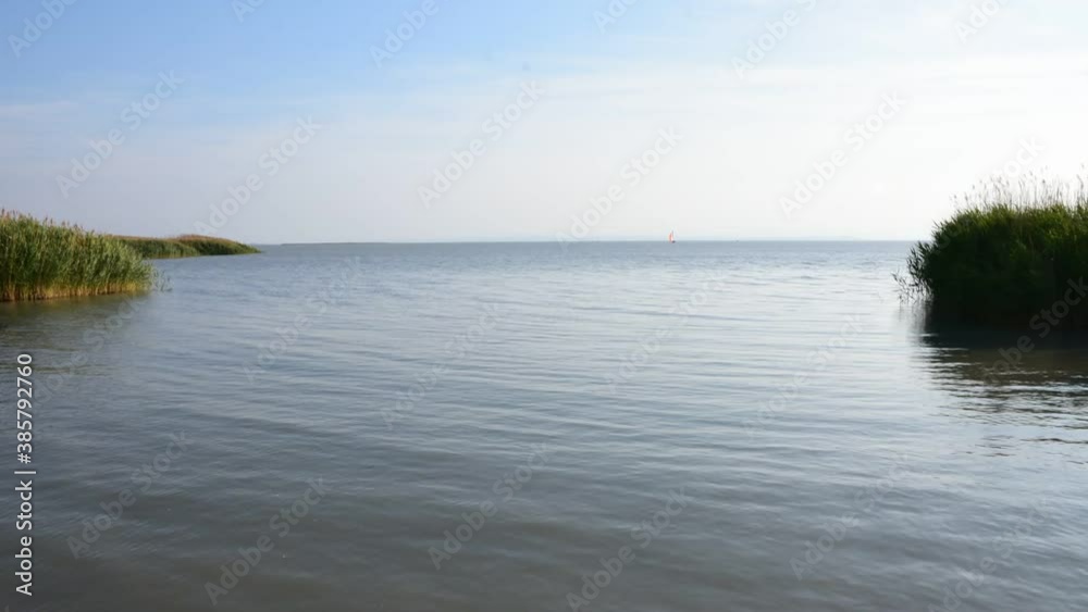 Time-lapse scenery at Lake Neusiedlersee in summer afternoon. Boats pass by in a sheltered bay with a belt of reeds in windy conditions.