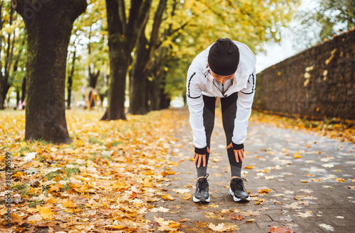 Fotografie Mid aged fit athletic woman dressed modern running clothes standing and bend over to stretch the back on the footway after jogging in the autumnal city park
