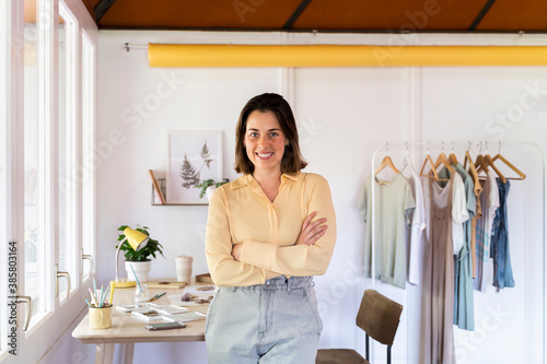 Smiling young female fashion designer with arms crossed leaning on table at clothing store photo