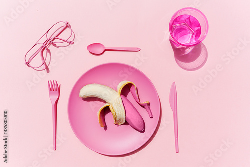 Studio shot of pink plastic tableware, glass of water, eyeglasses and pink peeled banana