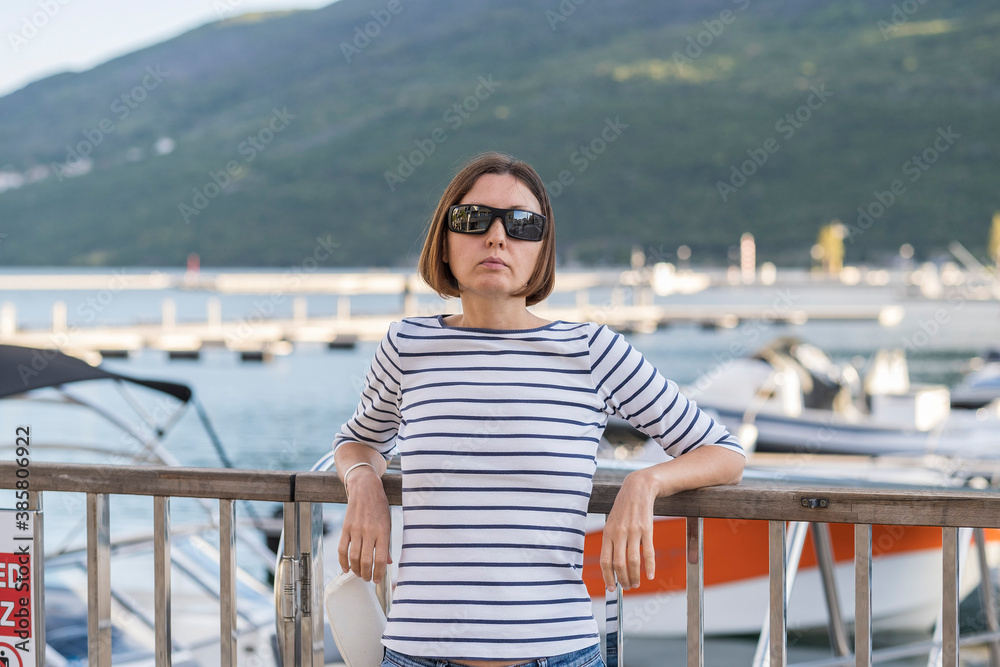 Young woman tourist with short brown hair in sunglasses and a white T-shirt with a blue strips is walking along the embankment along the sea. In the background, white boats and yachts are visible at t