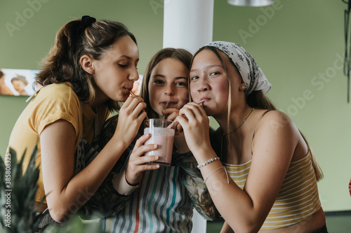 Teenage girls standing in kitchen sharing fresh fruit smoothie with drinking straws