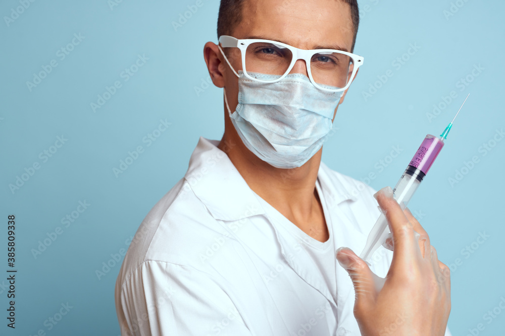 Doctor holds a syringe in his hand with a red liquid on a blue background medical gown and a protective mask Doctor holds a syringe in his hand with a red liquid on a blue background medical gown and a protective mask