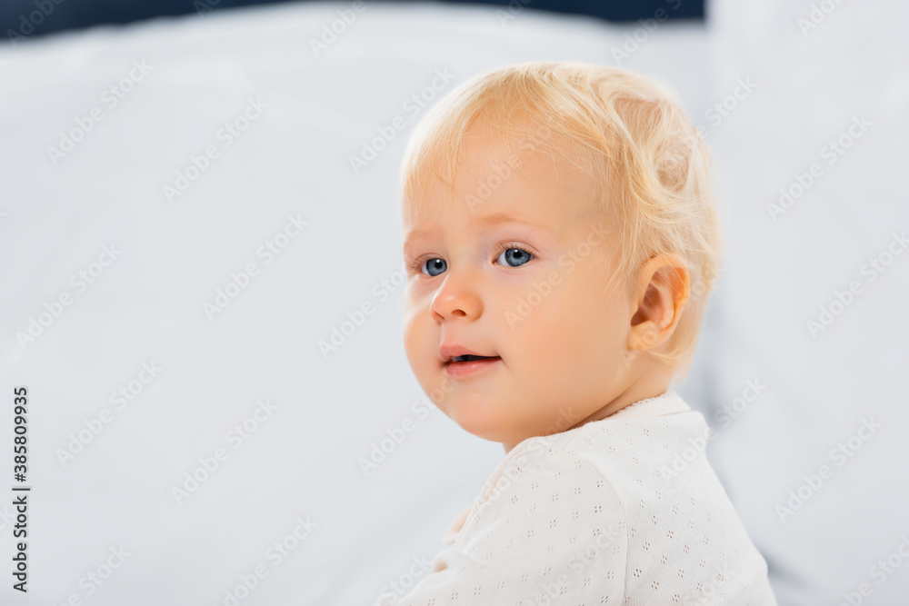 Selective focus of blonde toddler boy looking away on bed on white