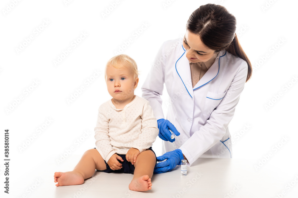 Doctor holding syringe with vaccine near toddler boy on table isolated on white Doctor holding syringe with vaccine near toddler boy on table isolated on white