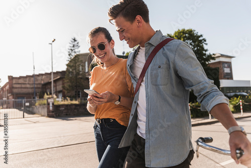 Smiling young couple looking at mobile phone while walking on street in city